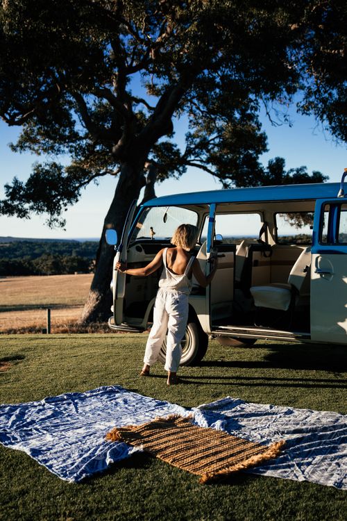 Back view of unrecognizable female traveler standing near opened doors of retro minibus while preparing camping with colorful blankets against picturesque nature background