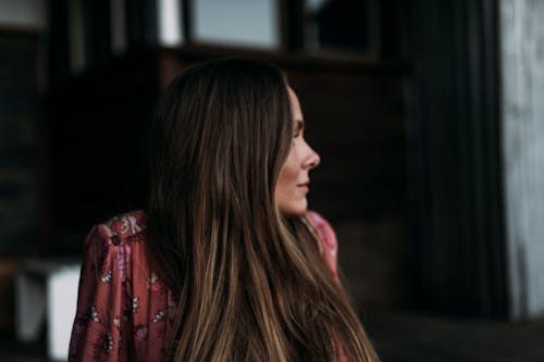 Free Side view of young woman with long dark hair wearing trendy long sleeved dress looking away on blurred background Stock Photo