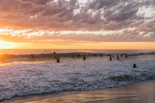 Silhouettes of surfers spending time in ocean with foamy waves splashing towards sandy shore at bright sunset on horizon