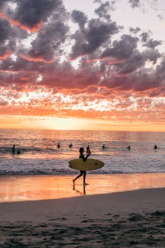 Unrecognizable people in swimwear surfing and swimming on sandy summer beach in calm sea at sundown