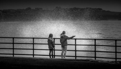 Black and white back view of unrecognizable tourists contemplating sea with splattering water while standing on fenced dock