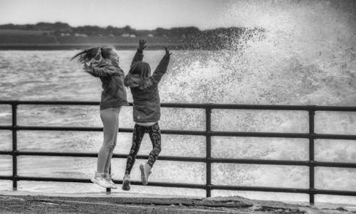Black and white back view of joyful anonymous travelers having fun with raised arms on fenced dock while enjoying stormy sea with splattering water