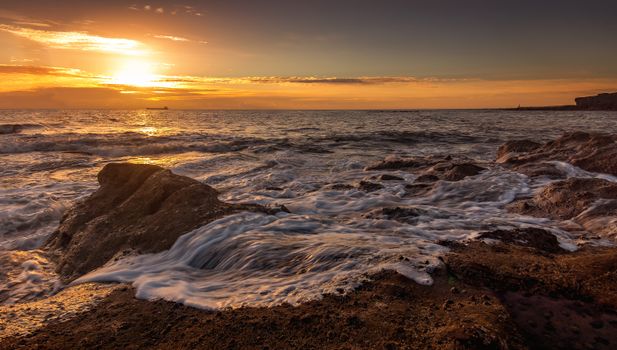 Scenery view of rapid ocean water fluid near rocky formations under sky with glowing sun in evening