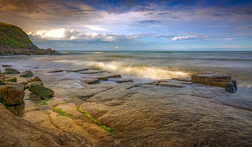 Wavy ocean with pure water near stones and mountain under bright cloudy sky with horizon