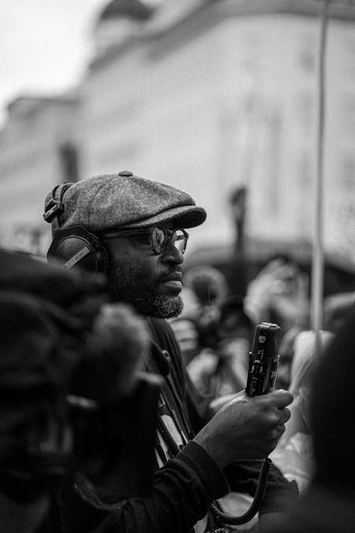 Black and white side view of African American male correspondent in eyeglasses and headphones with microphone during demonstration in city