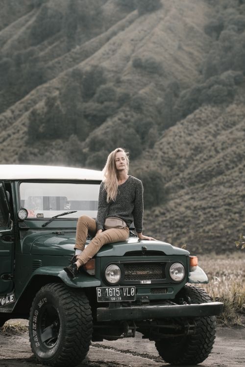Free Blonde woman sitting on a Jeep amidst a picturesque mountain backdrop, exuding adventure and freedom. Stock Photo