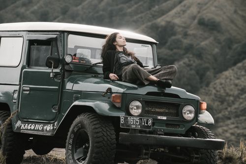 Free A woman relaxes on the hood of a green SUV, enjoying the serene natural surroundings of hills and grass. Stock Photo
