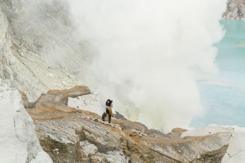 Free Person exploring a rocky volcanic crater with sulfuric fumes rising near a turquoise lake. Stock Photo