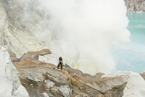 Free Woman stands amidst rocky volcanic terrain near steaming crater edge. Adventure concept. Stock Photo