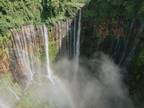 Aerial view of a stunning waterfall surrounded by lush greenery, with mist and a rainbow.