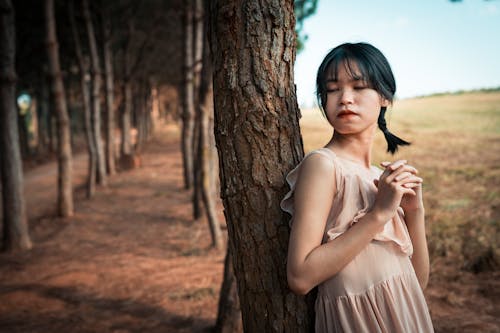 Dreamy tender Asian female in light dress closing eyes and leaning on tree trunk in sunny day