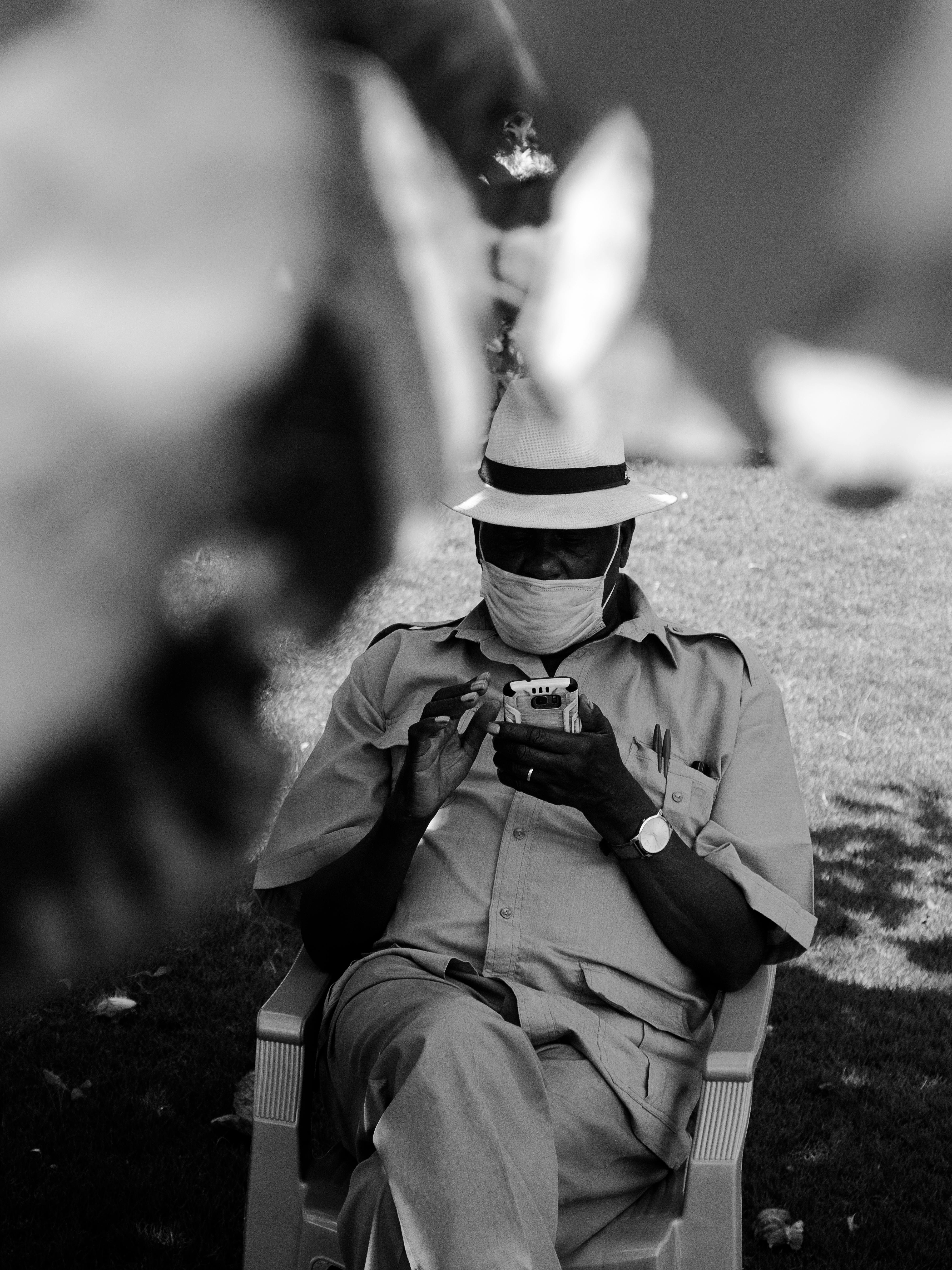 Black and white of unrecognizable African American middle aged male in casual clothes and face mask resting on chair in park and browsing smartphone