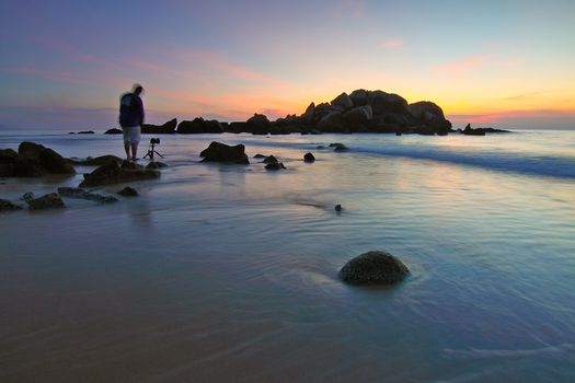 A lone photographer captures the serene seascape at sunset on a rocky beach.