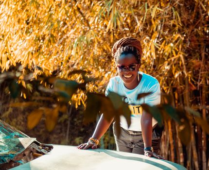 A joyful woman with braided hair poses stylishly in front of a car, surrounded by bamboo trees, wearing sunglasses, and a white t-shirt.