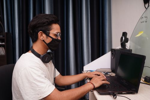 Free A young man wearing a mask works on his laptop at a home office desk, surrounded by tech accessories. Stock Photo