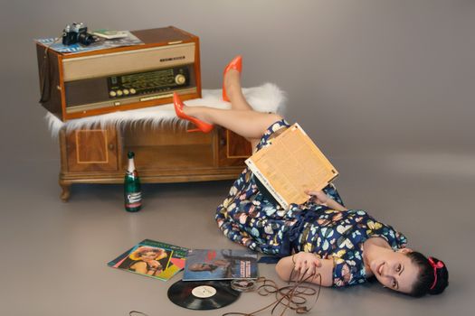 Full body of positive young female in old fashioned outfit lying on floor near vinyl records and retro audio receiver in gray studio