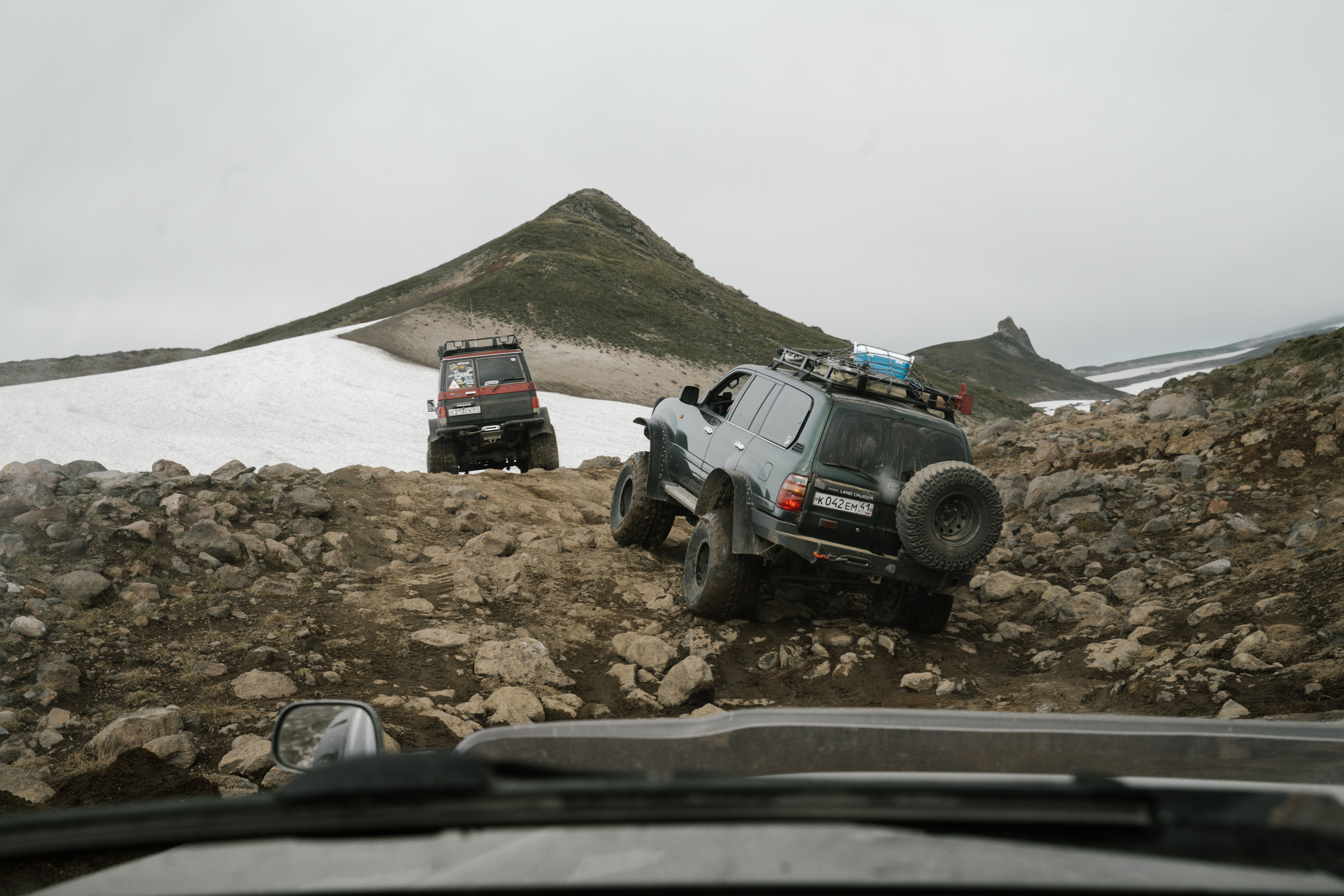 Two jeeps maneuvering a rocky, remote mountain path under an overcast sky.