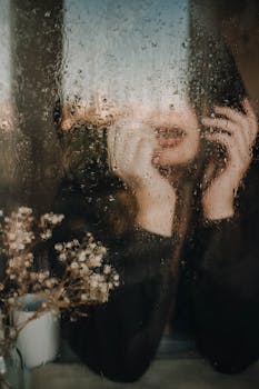 A contemplative woman leans against a rain-covered window with blurred reflections, conveying a moody atmosphere.