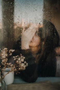 一名女子手持杯子，坐在布满雨痕的窗后，营造出一种忧郁而内省的氛围