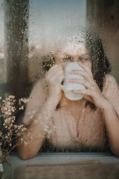 Free A woman enjoys a hot coffee while sitting behind a rain-covered window, creating a cozy and introspective mood. Stock Photo
