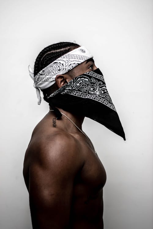 Side view of anonymous young shirtless black guy with Afro braids headband and bandana on face standing against white background