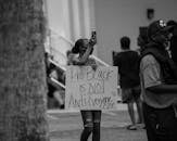 Grayscale Photo of Woman in Black Jacket Holding Smartphone Taking Photo of People