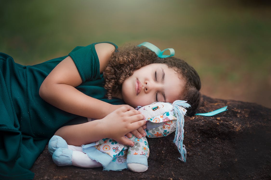 Free Adorable ethnic little girl with curly hair in dress hugging soft toy while sleeping on ground in park Stock Photo