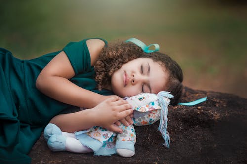 Free Adorable ethnic little girl with curly hair in dress hugging soft toy while sleeping on ground in park Stock Photo