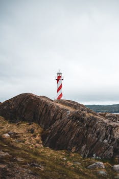 在阴沉的天空下，一座红白相间的灯塔矗立在崎岖的悬崖上，营造出孤独与宁静的氛围