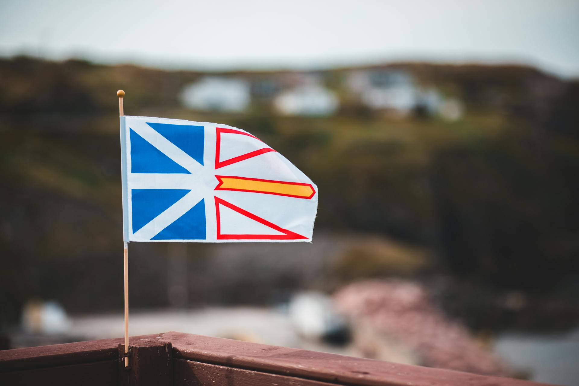 Le drapeau national de Terre-Neuve-et-Labrador est agité sur une clôture en bois contre une colonie côtière floue sur un terrain vallonné.