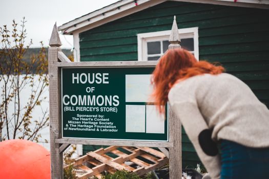 Back view unrecognizable female wearing casual clothes bending down and reading information on signpost with inscription House of Commons placed outside small rural house