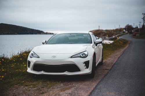 Modern white car parked on ground near asphalt road in village located on seashore on cloudy day