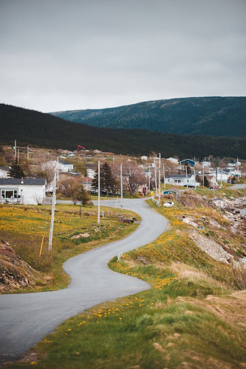 Empty curvy road going through grassy terrain in small village surrounded by green hills against overcast sky