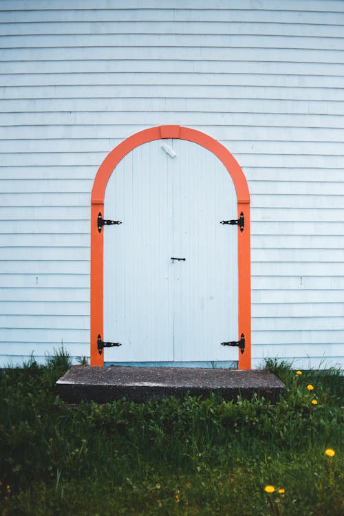 Arched white doorway with orange detail of simple wooden aged house located on grassy terrain in countryside