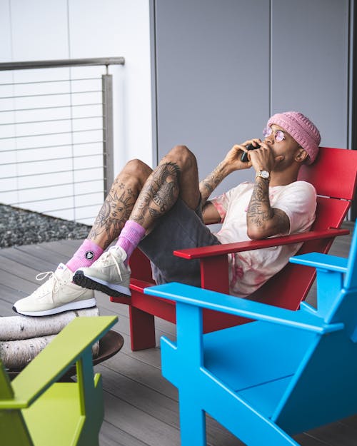 Side view of young ethnic tattooed guy in trendy outfit and sunglasses relaxing on wooden deckchair on terrace and talking on smartphone