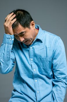 一名穿着牛仔衬衫的亚洲男子低着头，手放在头上，显得很担忧
