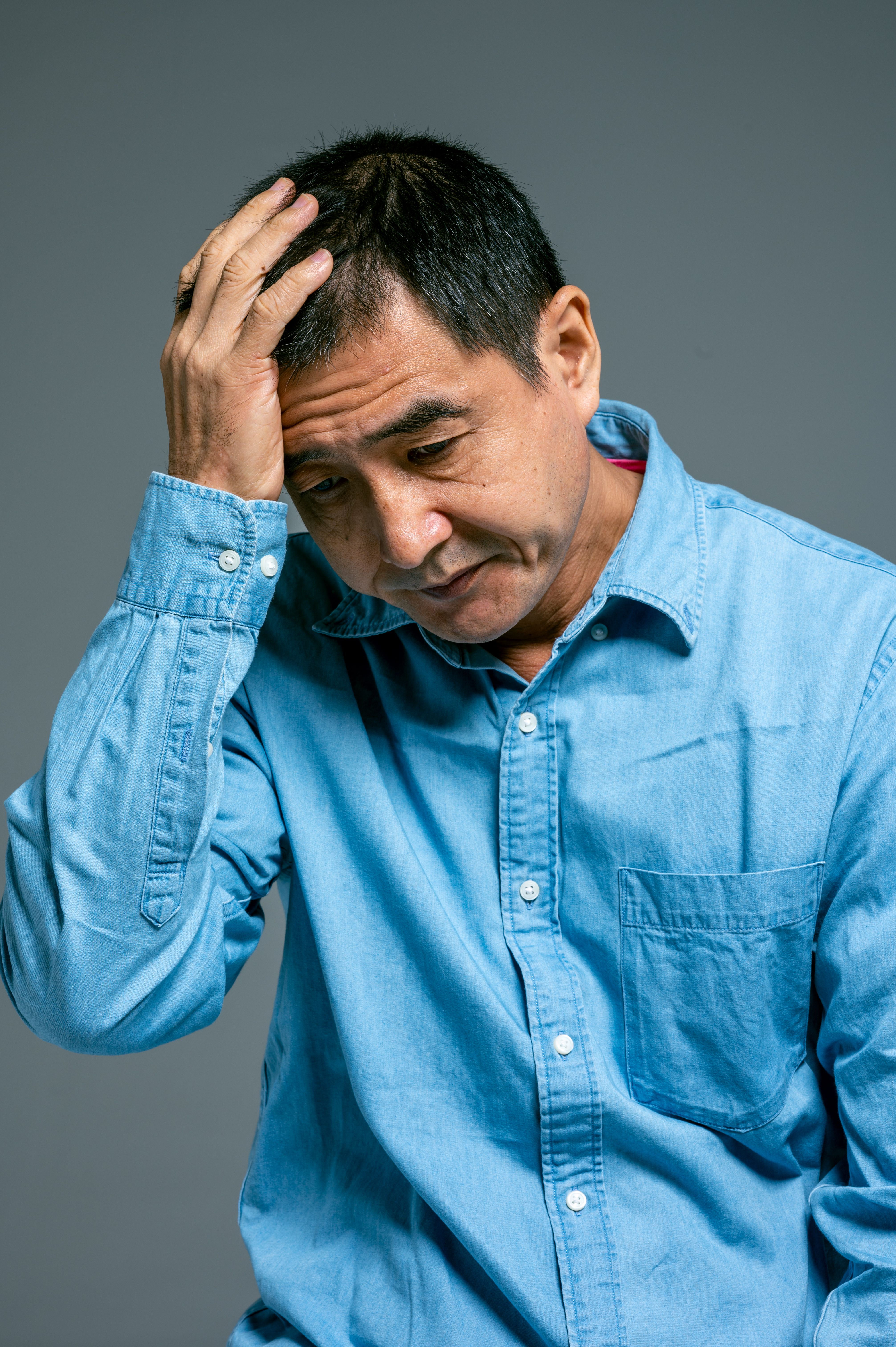 Mature man in blue denim shirt shows worry by holding head with hand. Emotional portrait.