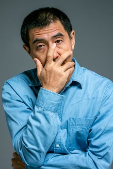 一名穿着蓝色牛仔衬衫的亚洲男子，手放在脸上，陷入沉思的特写