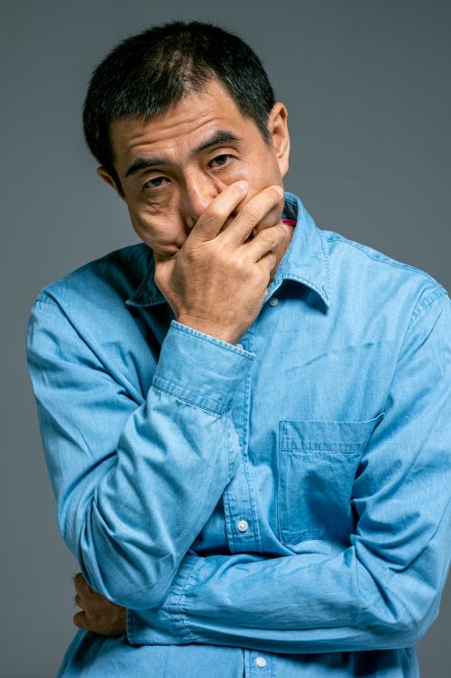 Free Thoughtful mature Asian man with hand on face, wearing blue denim shirt against gray background. Stock Photo
