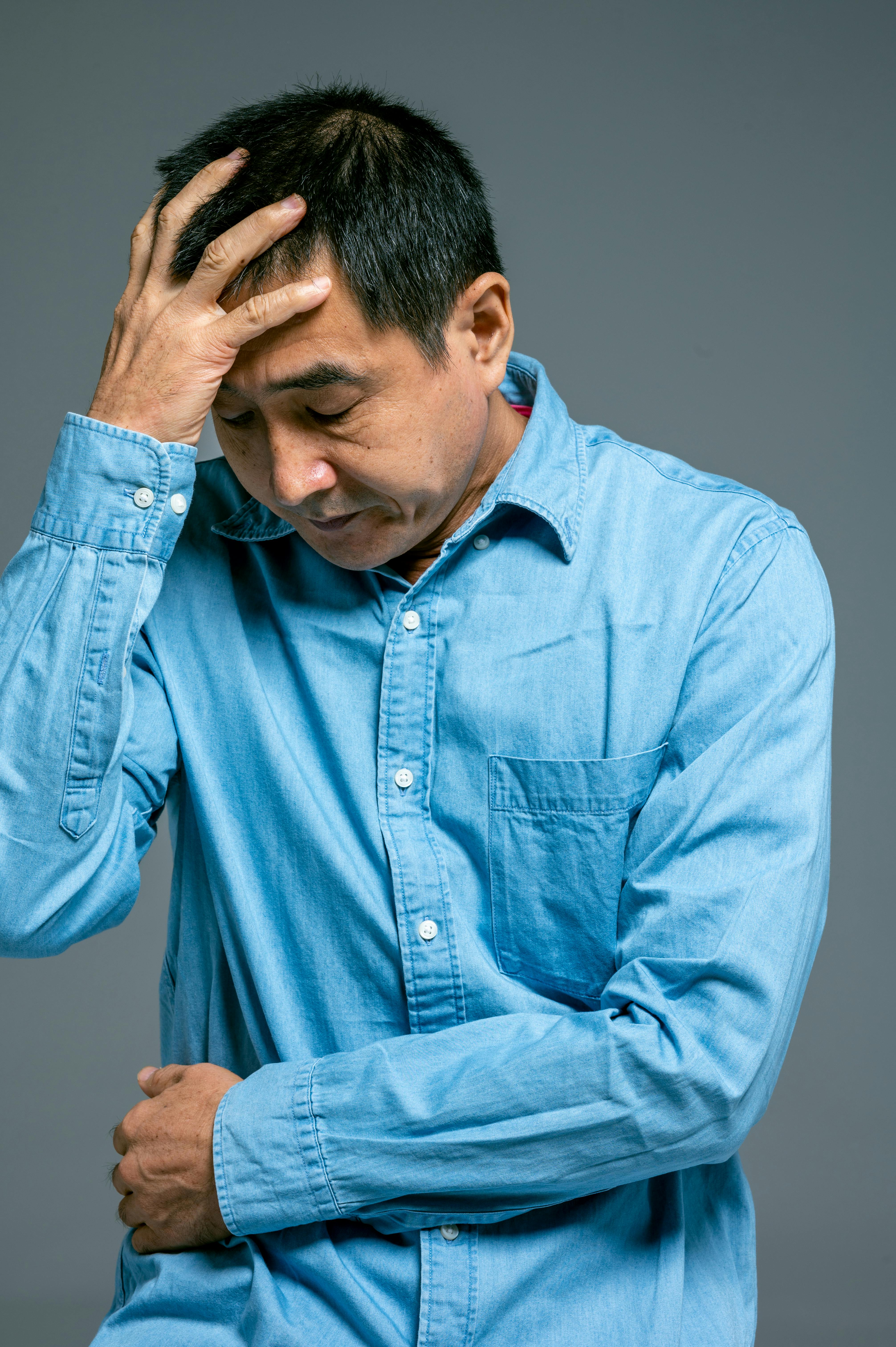 一位身穿蓝色牛仔衬衫的中年男子，手放在头上，表情若有所思