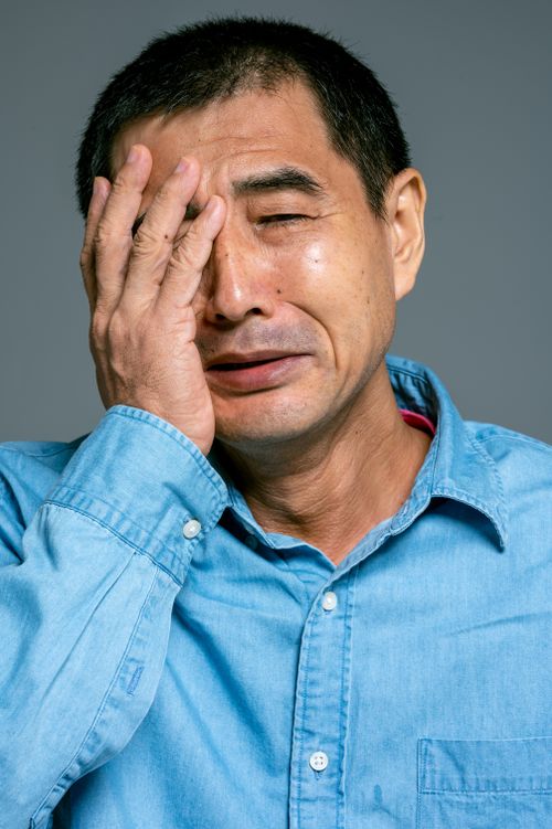 Free Close-up of an Asian man in a blue denim shirt expressing deep emotion with hand on face. Stock Photo