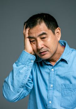 室内，一位穿着蓝色牛仔衬衫的亚洲男子手捂着脸，陷入沉思
