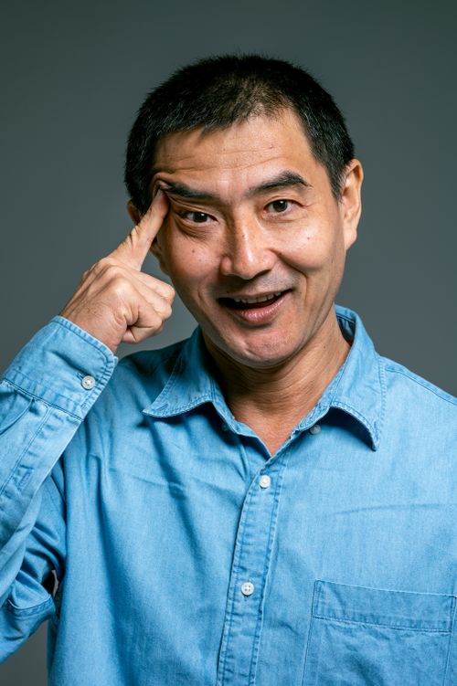 Free Asian man in denim shirt smiling and pointing to his temple indicating an idea or thought. Stock Photo