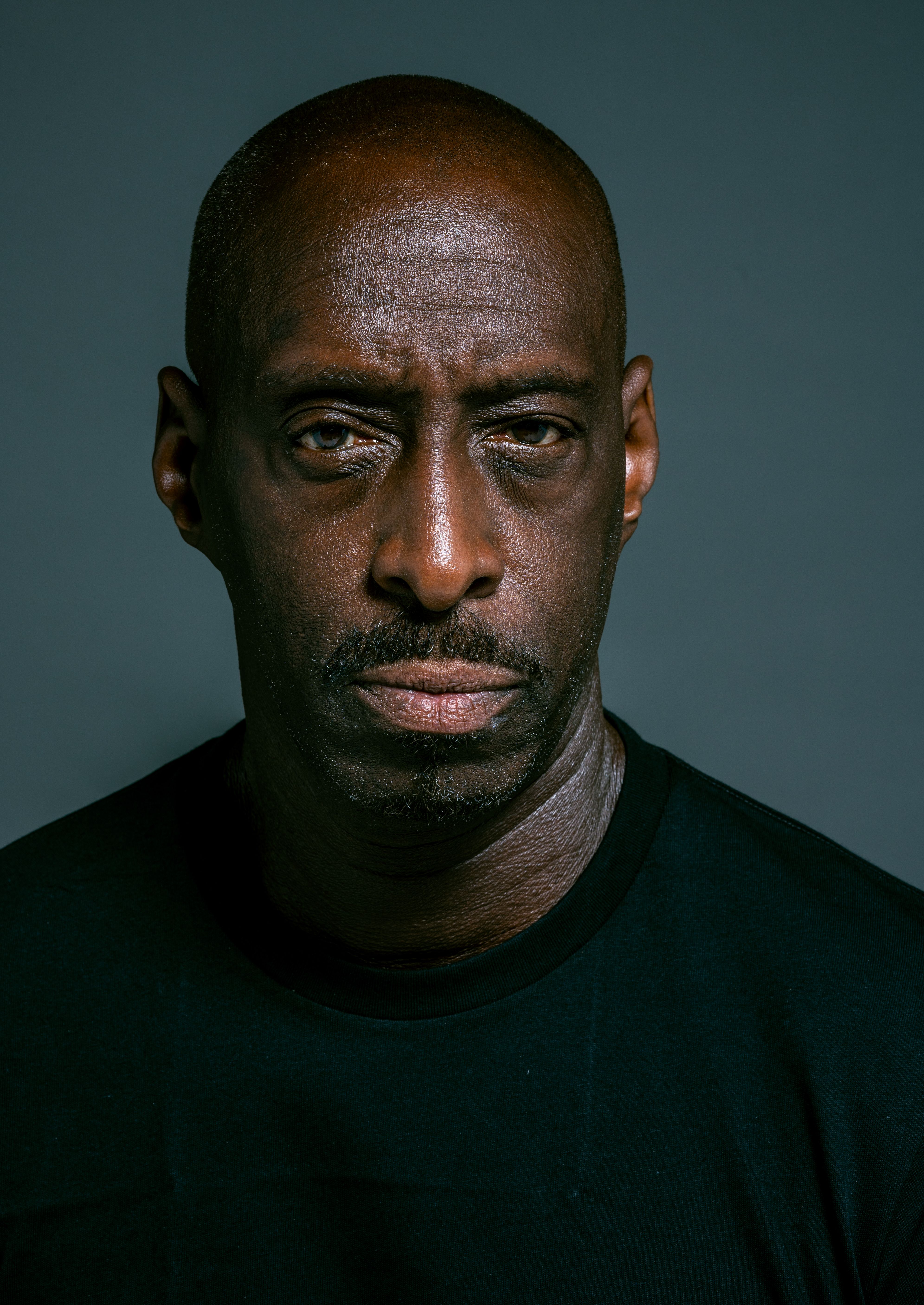 Close-up portrait of a serious man with a bald head in a black shirt against a dark background.
