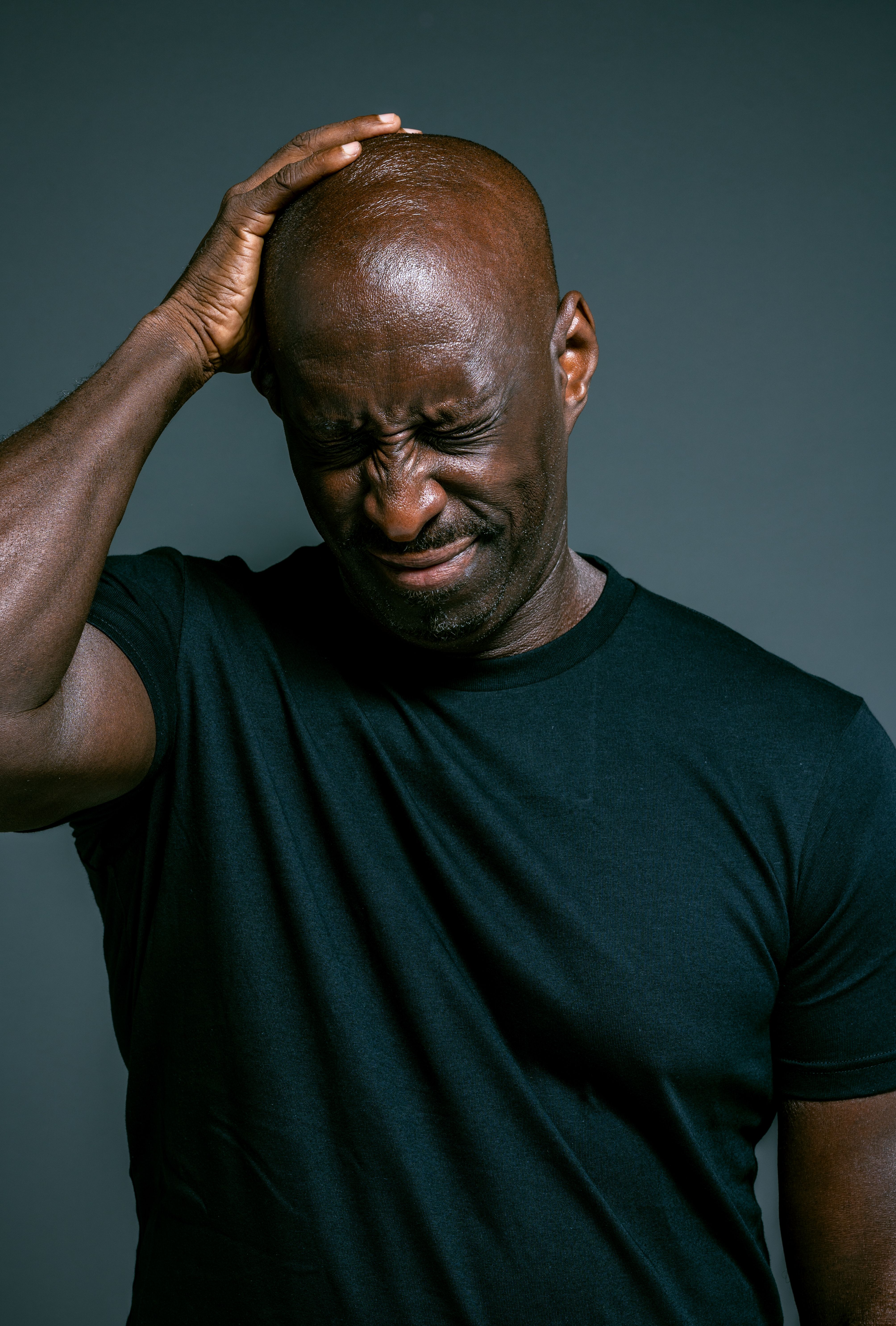 A man in a black shirt shows discomfort, touching his head against a gray background.