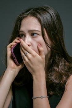 A concerned woman talking on the phone, covering her mouth with her hand, showing emotional distress.