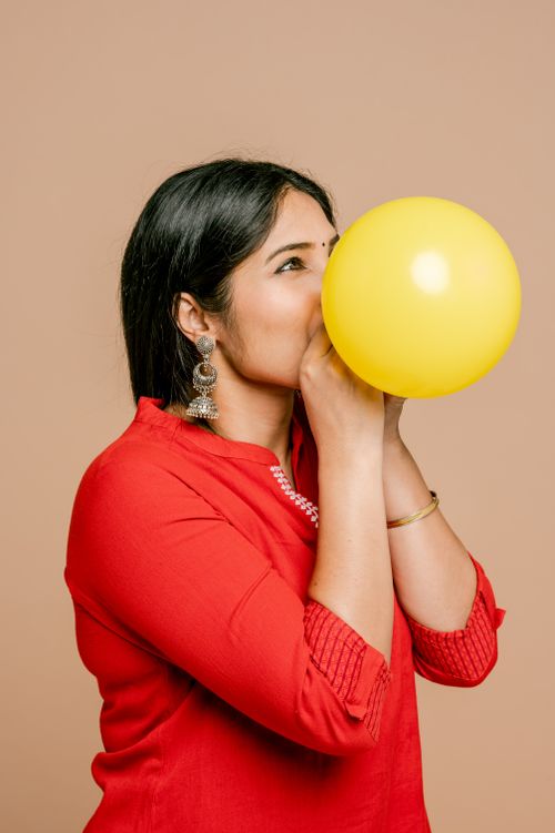Free A woman wearing a red dress blows a yellow balloon against a beige background. Stock Photo