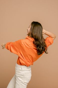 一位穿着橙色衬衫和白色裤子的年轻女子，在米色背景前快乐地跳舞