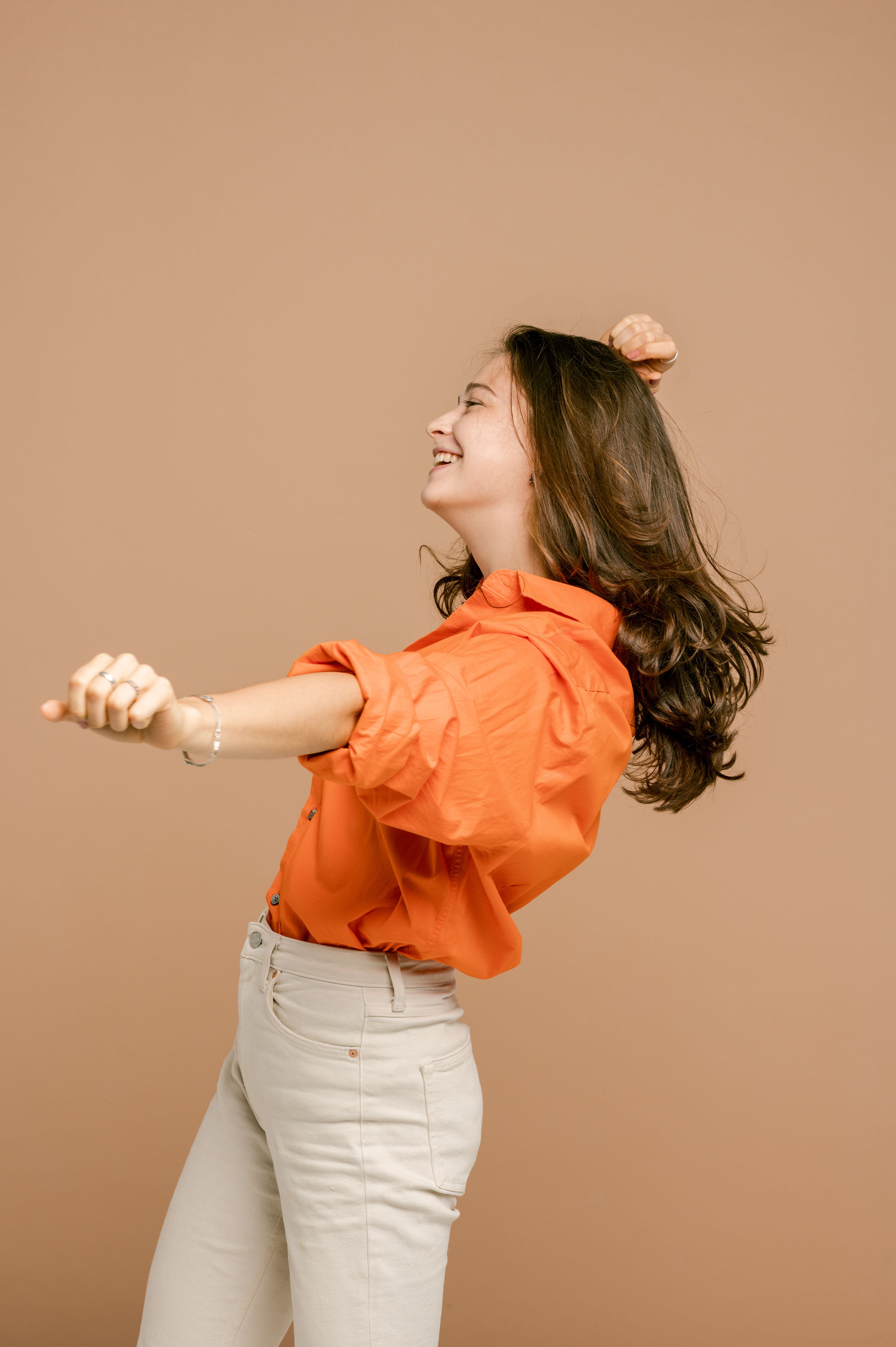 一名年轻女子身着鲜艳的橙色衬衫，在米色背景前快乐地跳舞、微笑