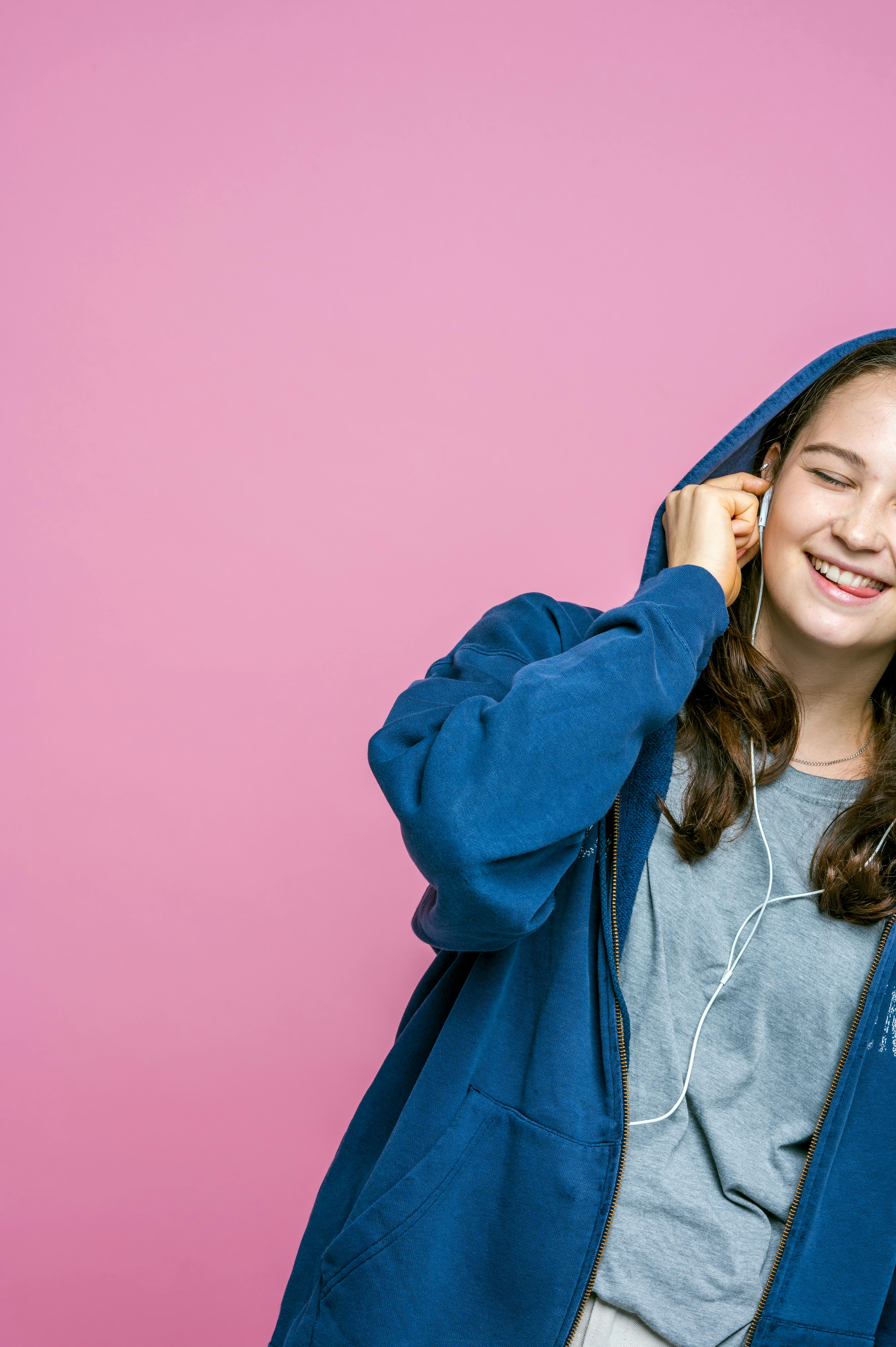 A young woman in a blue hoodie enjoys music with earphones against a pink background.